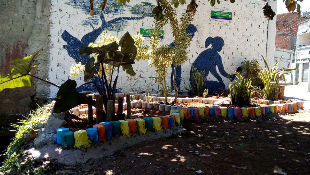 Ponto de revitalização na Avenida Coronel José Gladiador. no Morro Doce,No fundo há uma pintura no muro branco de um adulto e uma criança cuidando do jardim. Há plantações dentro de jardineiras coloridas.